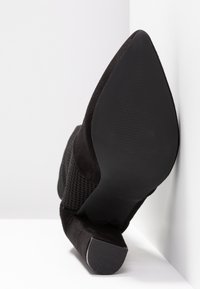 Bottine en daim noir à bout pointu avec tige en maille texturée et talon carré, photographiée de côté avec la semelle visible sur un fond blanc en angle.