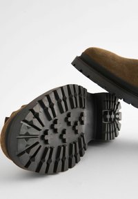 Brązowe zamszowe buty z wytrzymałą czarną gumową podeszwą, z głębokim bieżnikiem. Design obejmuje zaokrąglony czubek oraz teksturowane akcenty.