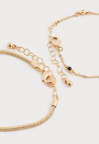 Dva zlaté řetízky na zápěstí s karabinkou, jeden s hladkým designem hadího článku a druhý s malým černým kamenem jako detailem.