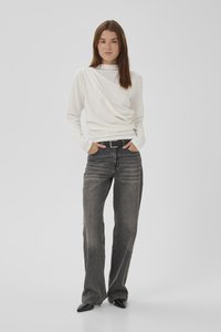 Femme debout, les mains dans les poches, portant un haut blanc à manches longues, une ceinture noire, un jean noir délavé et des chaussures noires pointues sur un fond uni.