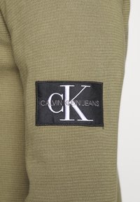 Olivgrön texturerad tyg med en svart rektangulär logotypapplikation som visar "CK" och "CALVIN KLEIN JEANS" i vit skrift.