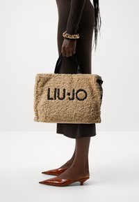 Beżowa torba tote z sztucznego futra z czarnym logo "LIU JO". Posiada czarne uchwyty, pudełkowaty kształt oraz teksturowaną powierzchnię. Spiczaste brązowe szpilki.