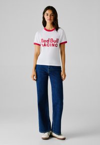 Tricou alb cu detalii roșii și inscripția "Red Bull RACING" imprimată cu litere îngroșate, asociat cu blugi întunecat flared și pantofi albi.