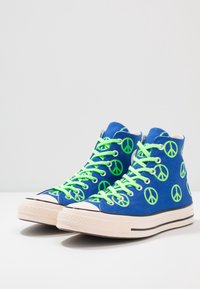 High-top sneakers in blue canvas with neon green peace sign prints, white rubber toe, and black-striped sole; features green laces.
