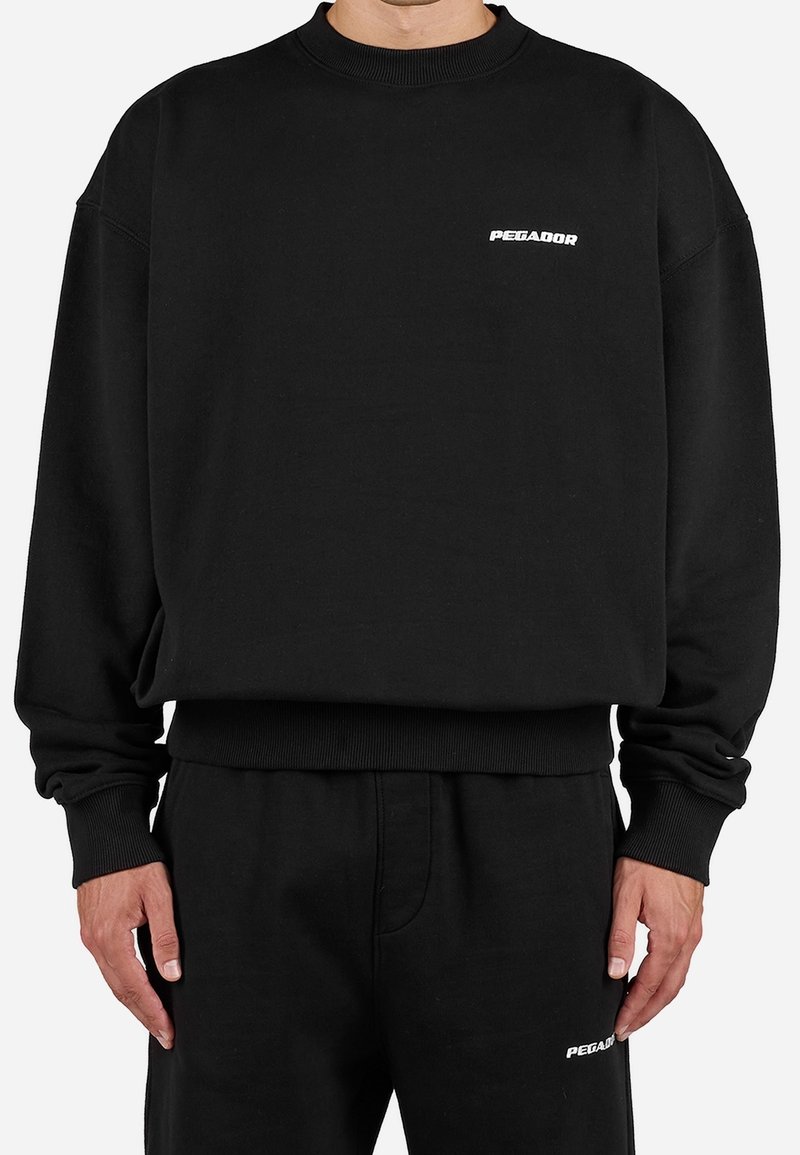Homme portant un sweat-shirt ample noir à col rond et un pantalon noir assorti, tous deux arborant un petit logo blanc "PEGADOR" sur le côté gauche.