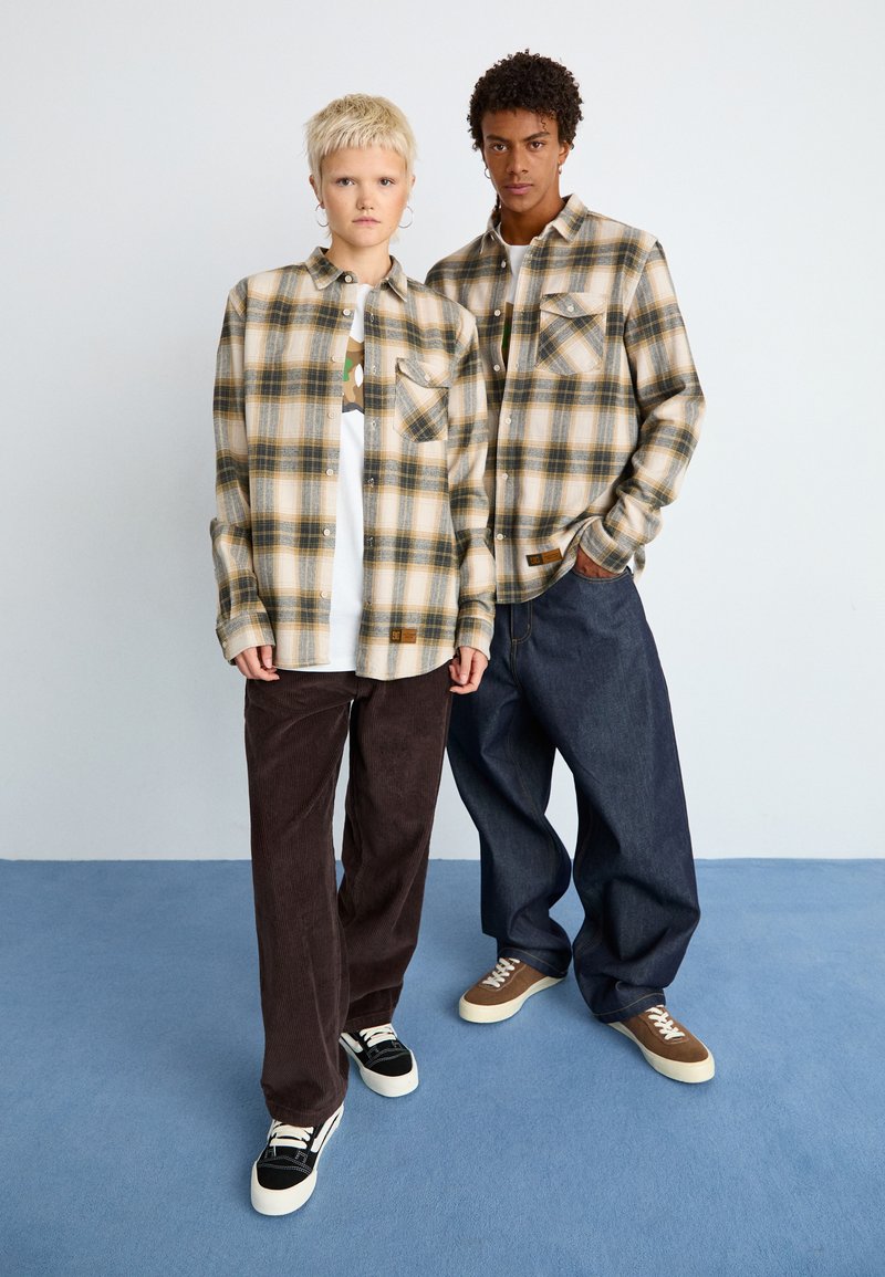 Dwie osoby noszą casualowe ubrania: jedna w beżowej koszuli w kratę i ciemnych spodniach, druga w podobnej koszuli z niebieskimi dżinsami. Obie mają na sobie sneakersy.