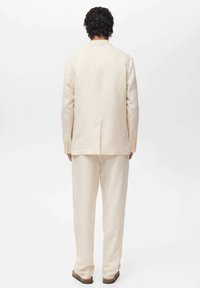 Lys beige linnedragt med en tætsiddende blazer og lige benbukser. Bagfra ses det minimalistiske design og de rene linjer.