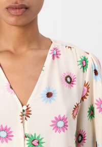 Bloemige blouse in crème met roze, blauwe, groene en bruine madeliefjes. Zachte stof met een v-halslijn en knoopdetail aan de voorkant.