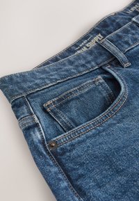 Gros plan sur un jean en denim bleu montrant la poche avant, les détails de couture, la boucle de ceinture et une partie de la ceinture avec un texte effacé.