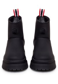 Botas negras hasta el tobillo con una parte superior sintética suave, puntera redondeada y suela de goma gruesa. Presentan un tirador de rayas rojas y negras en la parte trasera.