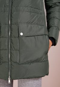 Zimní bunda v tmavě zelené barvě, s zipovým uzávěrem, prošívaným designem a přední kapsou se zapínáním na cvok. Hladká textura tkaniny.