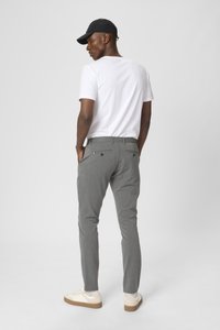 Grijze tailorpants met achterzakken, gecombineerd met een wit t-shirt en een zwarte pet. Sch footwear zijn witte sneakers met beige accenten.