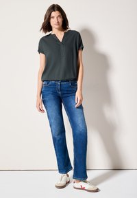 Mörkgrön kortärmad blus med V-ringning, i kombination med blå raka jeans och beige sneakers med röda accenter. Slät tygkvalitet.