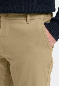 Chinos beige clair avec une texture lisse, présentant un devant plat, des poches latérales et un pull bleu marine contrastant partiellement visible.