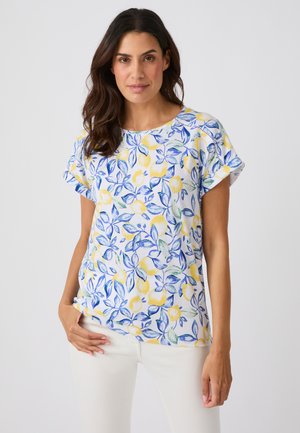 Femme aux cheveux foncés portant un haut blanc à manches courtes avec un motif floral et fruité bleu et jaune et un pantalon blanc sur un fond uni.