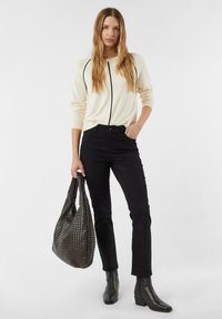 Frau mit langen Haaren, die einen hellen Pullover und schwarze Hose trägt, hält eine große gewebte schwarze Tasche in der rechten Hand und steht vor einem schlichten Hintergrund.