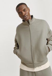 Pilkai zip-up džemperis su aukšta apykakle ir susiūtomis rankogaliais. Minkštas paviršius, laisvas įsipaišymas, su subtiliu logotipu ant krūtinės.