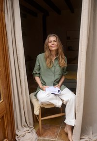 Femme aux cheveux longs portant une chemise verte et un pantalon blanc, assise pieds nus sur un tabouret en bois, tenant des papiers entre des rideaux beiges dans une pièce confortable.