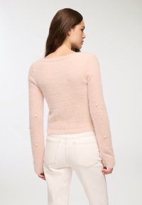 Jasno różowy, puszysty sweter z długimi rękawami, ozdobiony perłami na rękawach. Połączony z białymi dżinsami, widok z tyłu.