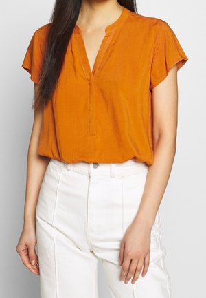 Femme portant un chemisier orange à manches courtes avec un col en V et un pantalon blanc taille haute, debout devant un fond uni.