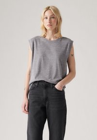 Blonde woman wearing sleeveless grey top and high-waisted black jeans, standing with one hand in pocket against plain background.
