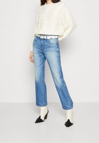 Pull en mailles blanches, jeans straight-leg en délavé clair, ceinture blanche à boucle. Le mannequin porte des bottes bicolores avec un bout pointu et un talon.