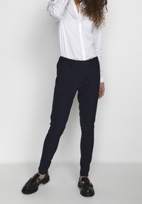 Marinblå slim-fit byxor med elastisk midja, slätt tyg och avsmalnande ben, kombinerat med en vit skjorta med knappar och svarta skor.