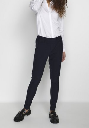 Pantaloni slim-fit blu navy con cintura elastica, tessuto liscio e gamba affusolata, abbinati a una camicia bianca e scarpe nere.