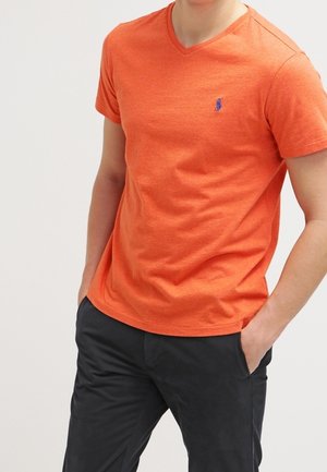 Mand iført en orange V-udskæring kortærmet skjorte med en lille blå logo på brystet og sorte bukser, hænderne i lommerne.