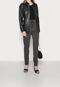 Černá kožená bunda ve stylu košile se dvěma předními kapsami, kombinovaná se šedými zúženými tepláky a černými otevřenými podpatky, držící malou kabelku.