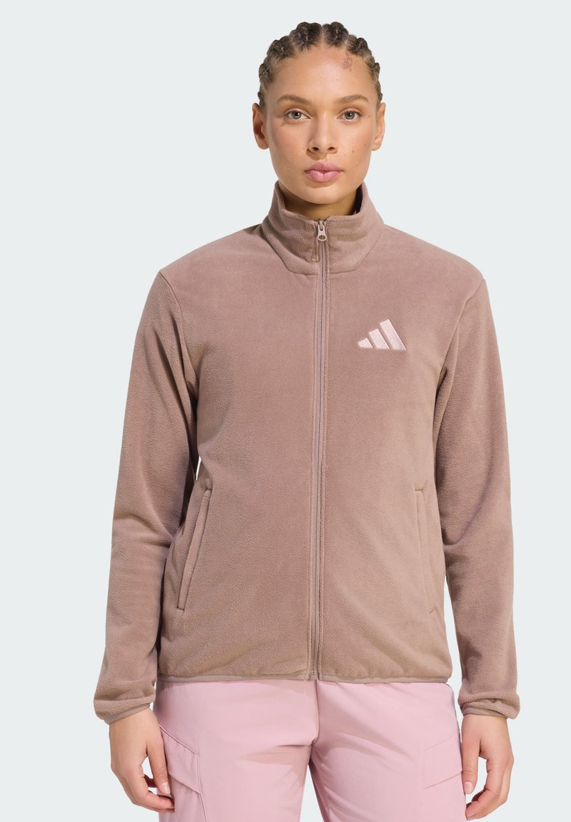 Γυναίκα που φοράει ένα ανοιχτό καφέ fleece μπουφάν με φερμουάρ και λογότυπο στο στήθος, συνδυασμένο με ανοιχτό ροζ παντελόνι, στέκεται μπροστά σε απλό φόντο.