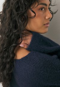 Kvinna med långt lockigt hår som bär en marinblå stickad tröja, håller kragen på sin axel, tittar åt höger mot en enfärgad bakgrund.