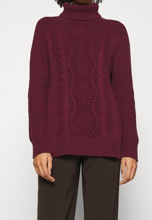 Pull col roulé bordeaux en maille torsadée avec motifs texturés, porté avec un pantalon marron foncé.