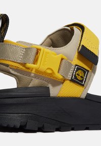 Beżowe sandały z żółtymi akcentami i czarną teksturowaną podeszwą. Posiadają żółty plastikowy klamrę i naszywkę z logo Timberland.