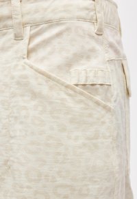 Κοντινό πλάνο μπεζ παντελονιών με διακριτικό λεοπάρ μοτίβο, που διαθέτουν λοξή μπροστινή τσέπη και μια μικρή θηλιά πάνω από αυτή.