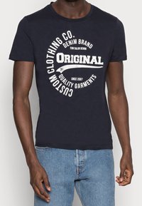 Camiseta de algodón azul marino con gráficos impresos en blanco y texto, diseño de cuello redondo y mangas cortas, combinada con jeans de mezclilla azul claro.