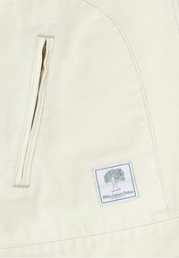 Veste en toile ivoire avec une poche zippée sur le côté, ornée d'une étiquette blanche avec un logo d'arbre vert et le texte "Belivs Originals Clothing".