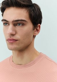 Lyserød tekstureret T-shirt med rund hals, der har korte ærmer og et subtilt mønster. Modellen har kort, mørkt hår og lys hud.