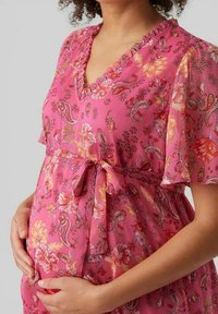 Robe de maternité fleurie rose avec un décolleté volanté et des manches courtes et légères. Présente une ceinture à nouer à la taille et une texture de tissu à motifs.