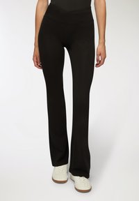 Pantaloni yoga neri a palazzo con una cintura larga e incrociata. Realizzati in un tessuto liscio e elastico. Indossati con sneakers bianche.