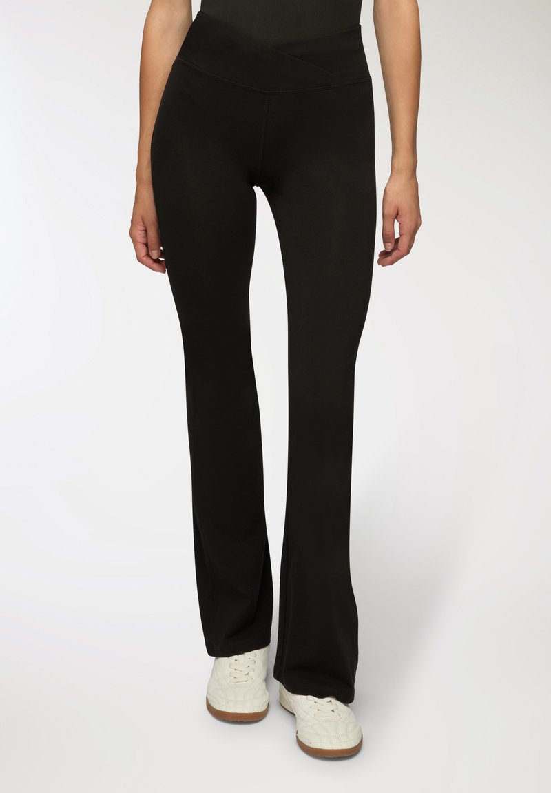 Pantaloni yoga neri a palazzo con una cintura larga e incrociata. Realizzati in un tessuto liscio e elastico. Indossati con sneakers bianche.