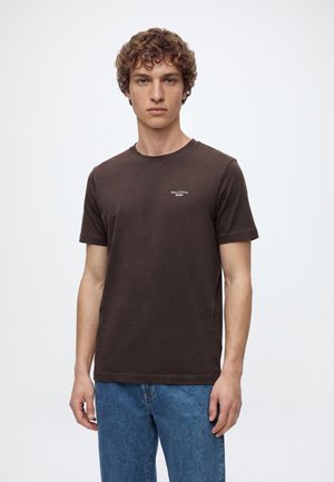 Joven con cabello rizado que lleva una camiseta marrón oscuro de Marc O'Polo y vaqueros azules, de pie frente a un fondo claro y sencillo.