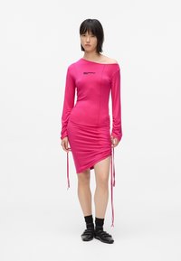 Γυναίκα φορώντας ένα φωτεινό ροζ φόρεμα με ρυάκωση και χωρίς ώμους, μαύρες κάλτσες και μαύρα παπούτσια, στέκεται μπροστά σε έναν απλό λευκό φόντο.