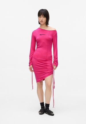 Vrouw in een felroze off-shoulder gerimpelde jurk, zwarte sokken en zwarte schoenen, staand tegen een plain witte achtergrond.