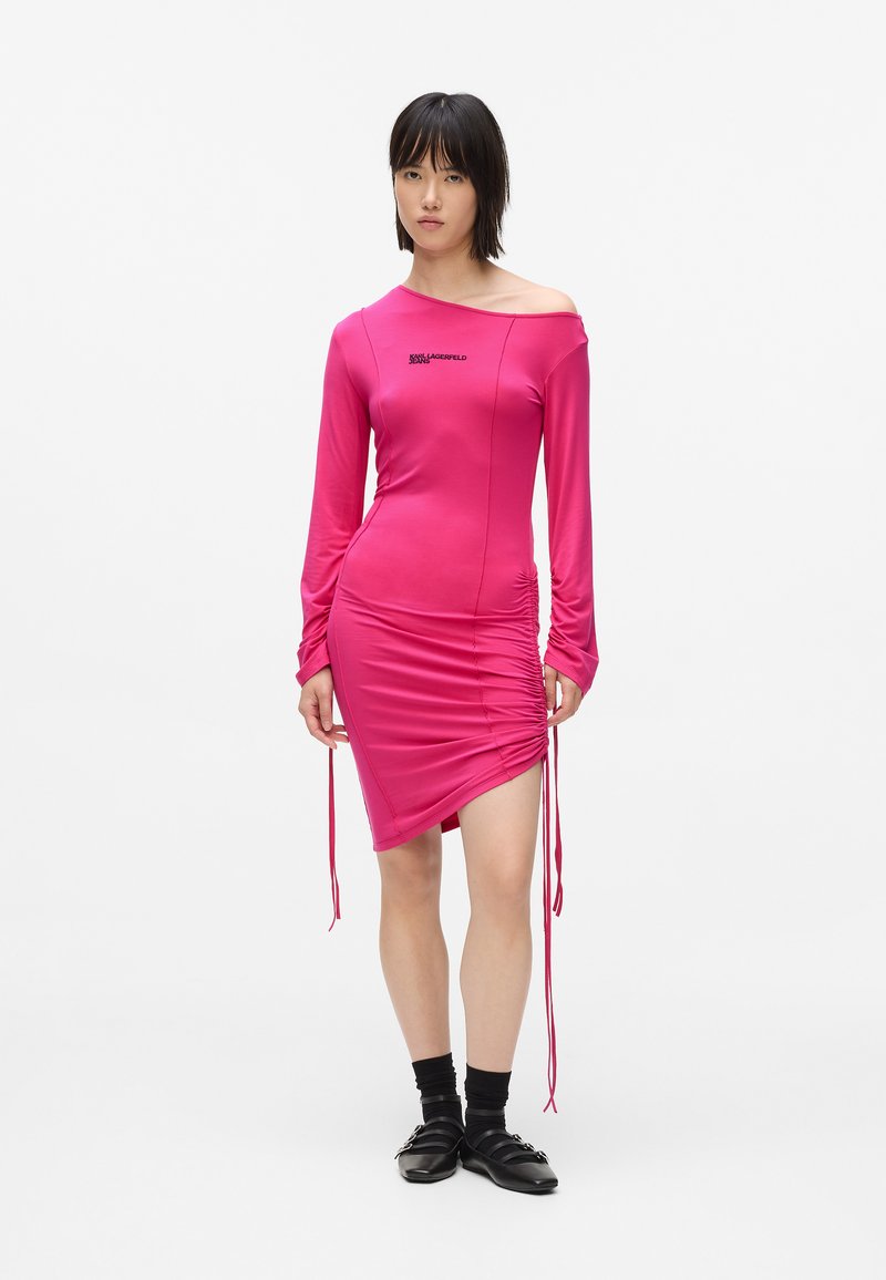 Γυναίκα φορώντας ένα φωτεινό ροζ φόρεμα με ρυ�άκωση και χωρίς ώμους, μαύρες κάλτσες και μαύρα παπούτσια, στέκεται μπροστά σε έναν απλό λευκό φόντο.