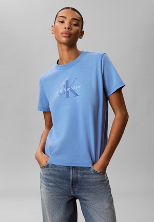 Vrouw met handen in haar zakken, draagt een lichtblauw Calvin Klein logo T-shirt en vervaagde blauwe spijkerbroek, staand tegen een effen grijze achtergrond.
