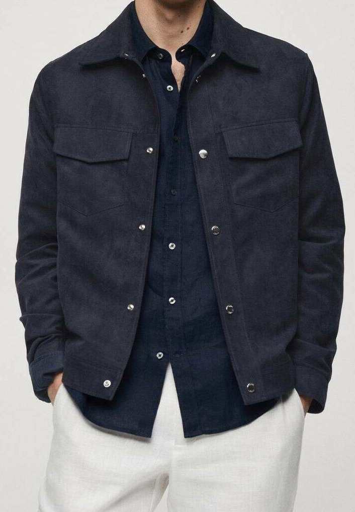 Tmavě námořnická semišová bunda s límcem, dvěma náprsními kapsami, druky a hladkou texturou, vrstvená přes tmavé tričko a bílé kalhoty.