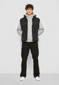 Sort puffer vest med hætte, grå hoodie under, parret med sorte cargo bukser og hvide sneakers. Har quiltet tekstur og lommer.