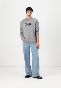 Felpa grigia con la scritta "PARIS", abbinata a jeans a gamba larga di un leggero blu. Modella indossa scarpe nere, in piedi su uno sfondo bianco.