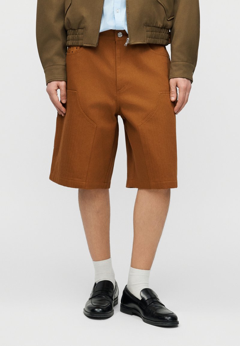 Άνδρας που φοράει καφέ σορτς μέχρι το γόνατο, λευκές κάλτσες, μαύρα επίσημα παπούτσια, ανοιχτό μπλε πουκάμισο και ελιάς πράσινο μπουφάν με ελαστική ραφή στο τελείωμα.